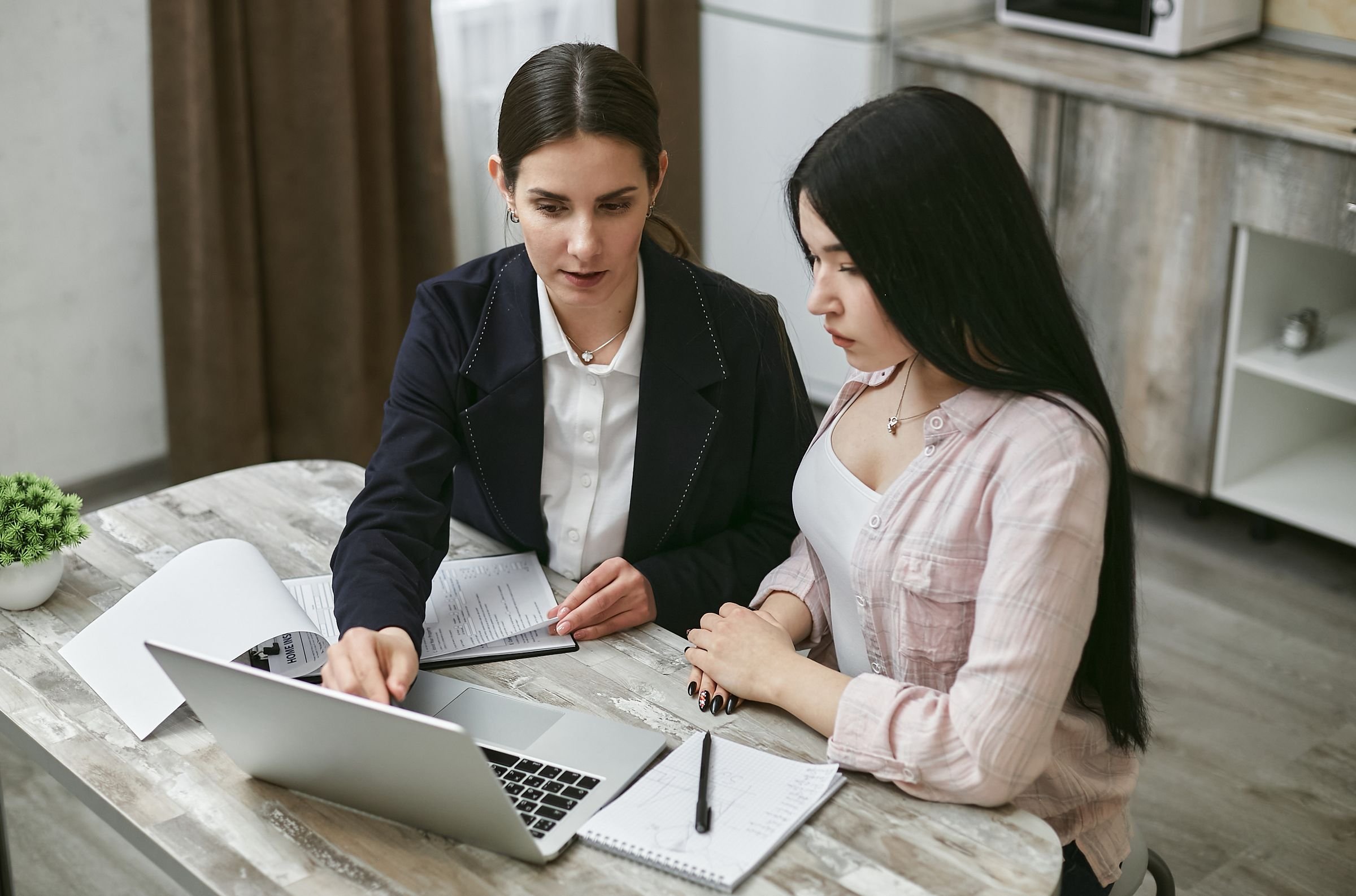 An advisor sitting beside a business owner at her kitchen table, walking her through a document on a laptop