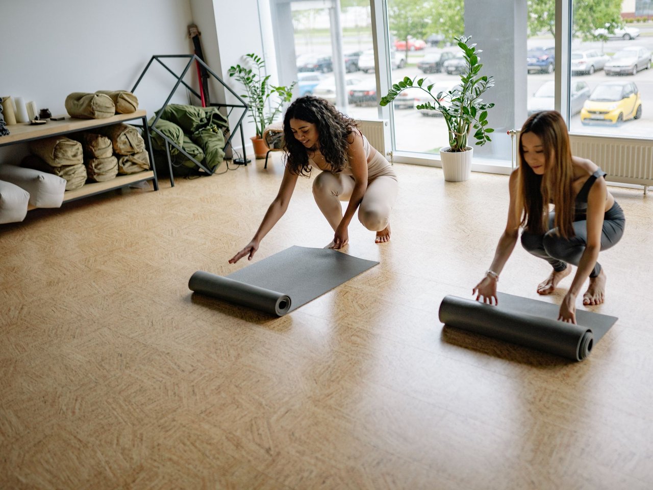 Two women unrolling yoga mats in a bright, sunlit studio.