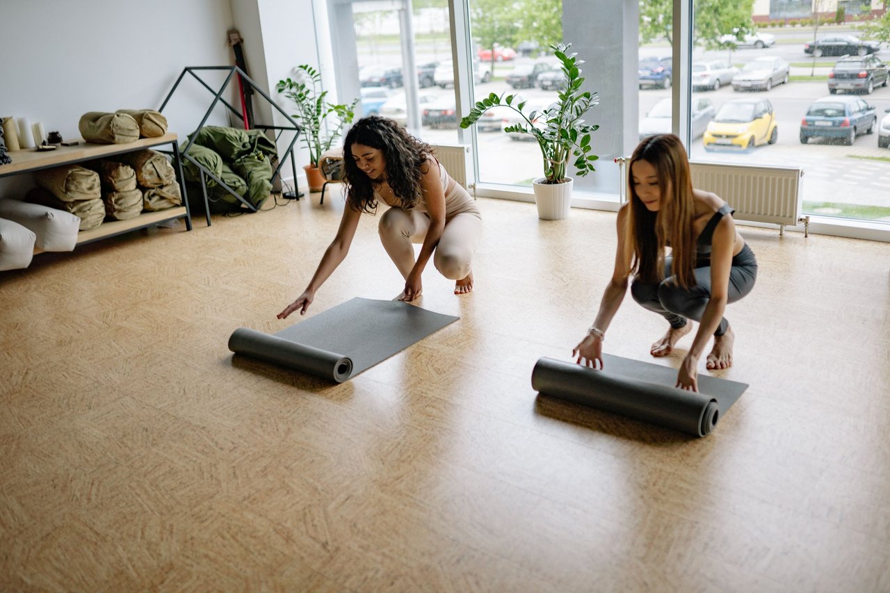 Two women unrolling yoga mats in a bright, sunlit studio.